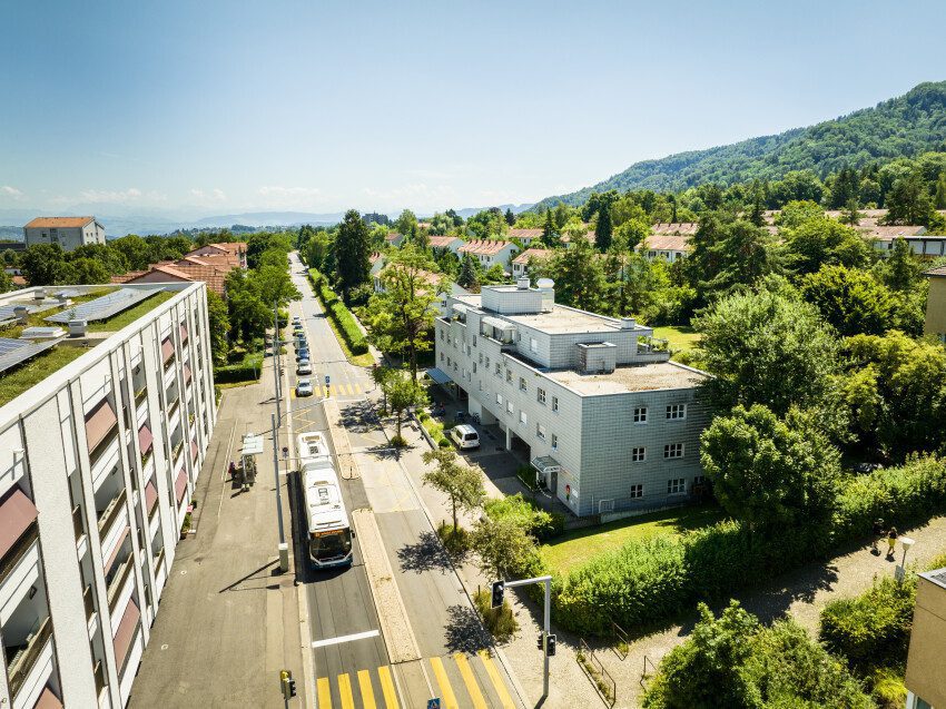 Luftaufnahme des Mehrfamilienhauses der Siedlung Verwaltungsgebäude (Etappe 21), davor die Schweighofstrasse mit einem Bus, gegenüber das Quartierzentrum Friesenberg