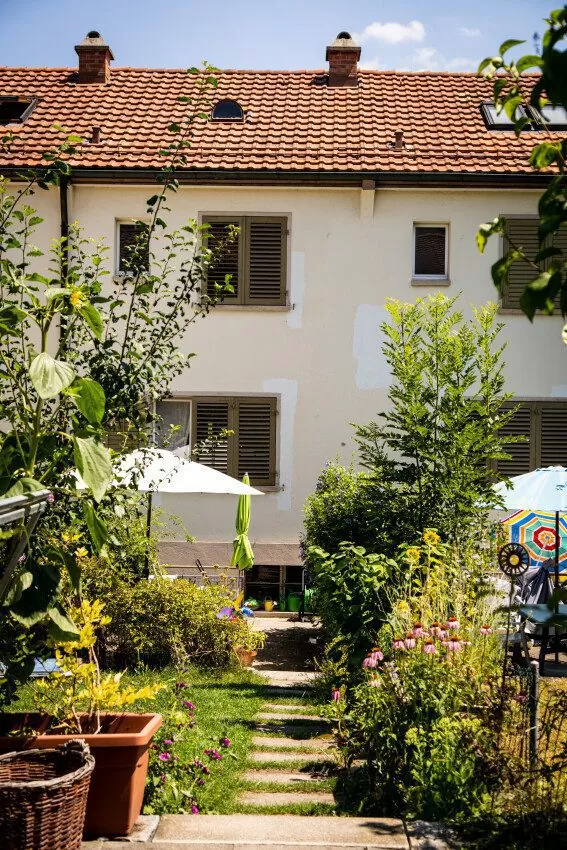 Gartenweg eingerahmt von Pflanzen und Töpfen, ein Sonnenschirm, dahinter ein Reihen-Einfamilienhaus der FGZ-Siedlung Schweighofstrasse Süd (Etappe 9)